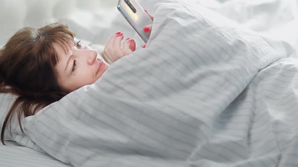 Sleepy Brunette Woman Lies on the Bed and Uses the Phone in the Morning Read Some Information