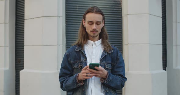 Hipster Young Man with Earrings Looking Aside While Using His Smartphone. Bearded Millennial Guy alt