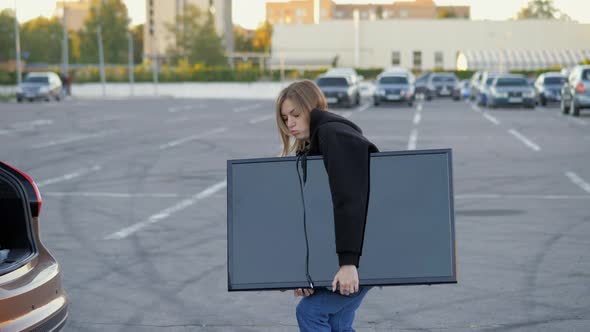 Woman Funny Carries a Modern Large TV in Hands Tries to Load It Into the Trunk of a Car on the alt
