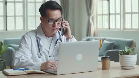 Asian Male Doctor Talking On Phone During Work On Laptop Computer At Home Office alt