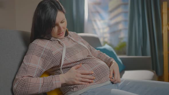 a Pregnant Woman is Sitting on the Couch and Experiencing Discomfort alt
