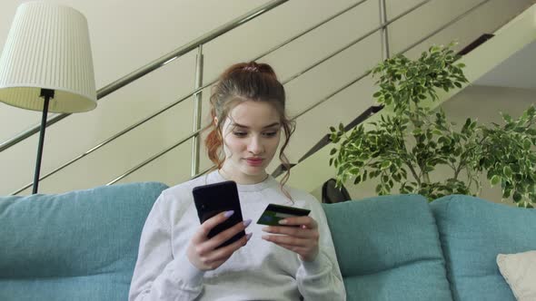 Pretty Girl is Making Online Payment Holding Bank Card at Home Sitting on Sofa and Smiling. alt