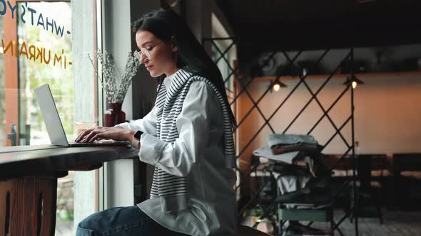 Confident young brunette woman in eyeglasses working on laptop alt