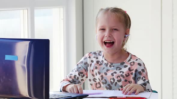 A little girl sits behind a laptop wearing headphones and laughs.