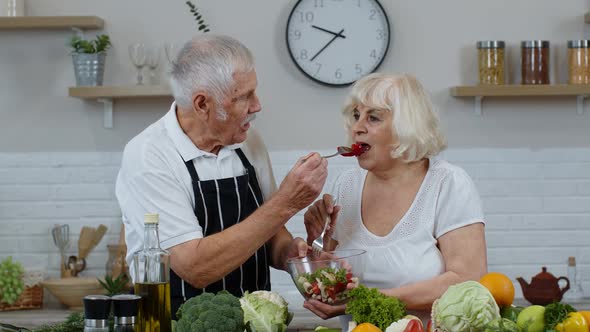 Senior Couple in Kitchen. Grandmother and Grandfather Feeding Each Other with Raw Vegetable Salad alt