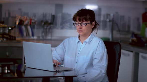 Woman with Glasses Sits at a Laptop in the Kitchen at Home Woman is Studying Information on the