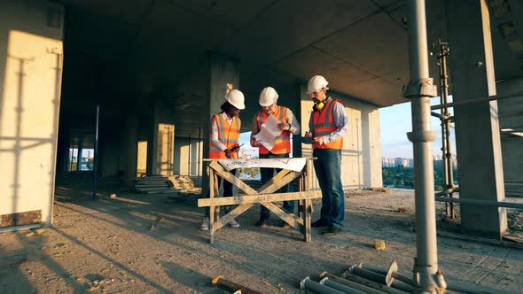 Three Construction Worker Look at a Blueprint on a Building Site. Multiethnic Engineers, Architects alt