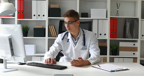 Young Doctor Holding Phone in Hand and Working at Computer