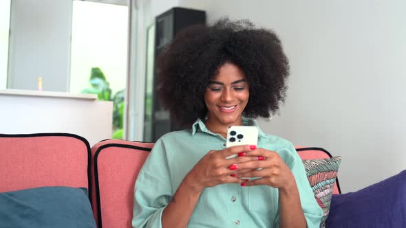 Young Smiling African Woman Sitting at the Sofa at Home and Using Smartphone