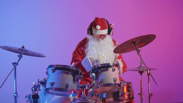 A Man Dressed As Santa Claus Plays a Drum Kit in a Dark Studio Against a Backdrop of Bright Purple alt