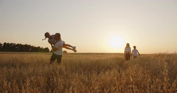 Family With Children Having Fun In The Field. Dad Circles The Girl In His Arms