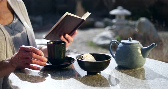 An elderly woman reading a book and drinking a cup of herbal tea with a bowl and a teapot in a Japan alt