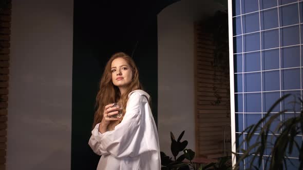 Red-Haired Girl In White Shirt Holds Glass Of Water In Her Apartment. alt