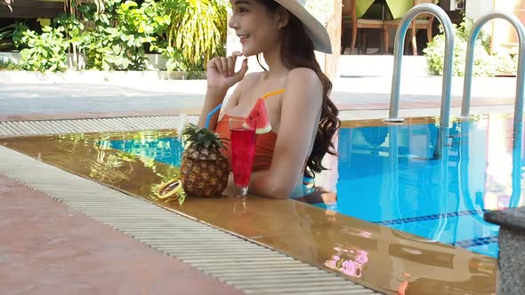 Summer Concept. Sexy asian woman in orange bikini with tropical drink in swimming pool. Tilt up shot