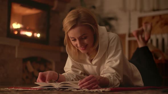 Pretty Blonde Girl Lying Near Fireplace and Reading a Book alt