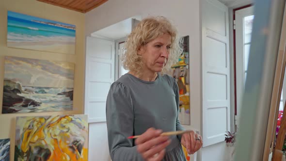 A Middleaged Woman Artist Paints a Picture in Front of a Window in an Art Workshop alt
