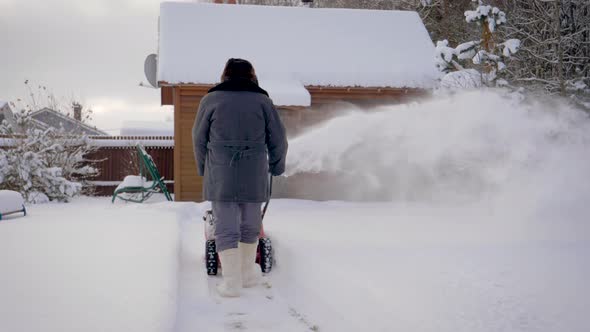 Man Cleans Snow With Snow Plow Background Of Wooden House In Winter alt