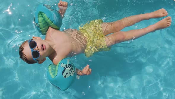 Relaxed Small boy wearing sunglasses floating on Swimming pool using colorful floaties alt