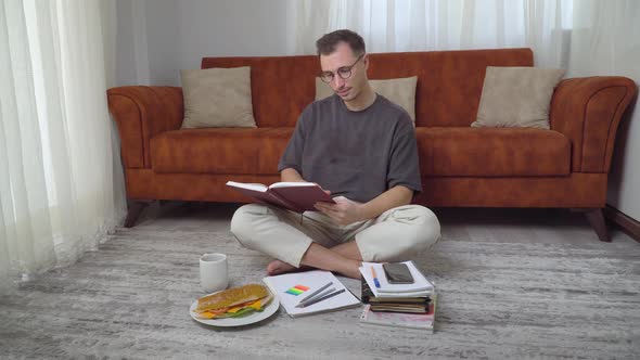 A Student Guy Reading a Book and Drinking Coffee at Home Sitting on the Floor