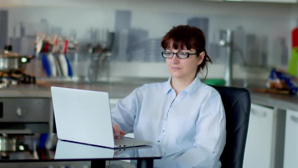 Brunette Woman Sits in the Kitchen and Works at a Laptop the Reflection From the Monitor in Glasses