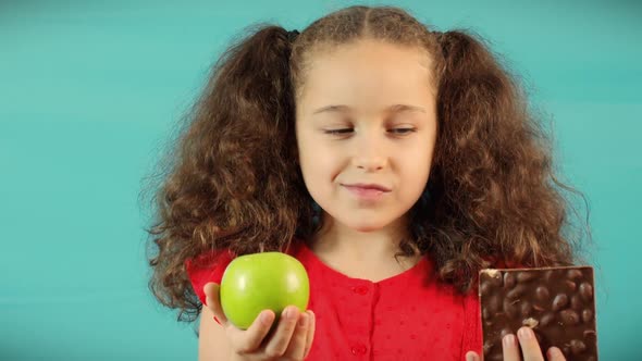 Little Girl Makes a Choice Between of Holding a Green Apple and Chocolate alt