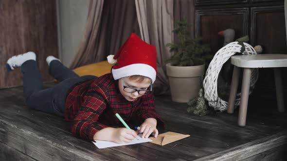 Boy in Christmas Hat and Glasses Lies on Floor and Writes Letter to Santa Claus alt