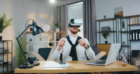 Man in Virtual Reality Headset Sitting at the Table in Home Office and Working on Imaginary Screen alt