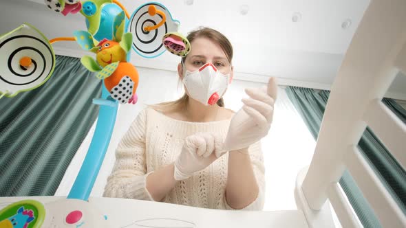 Young Mother Wearing Protective Medical Mask an Rubber Gloves Looking at Her Baby Lying in Cradle alt