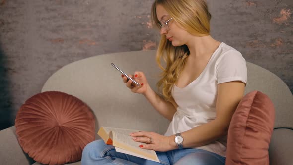 Young Woman with Book on Lap Texting Using Mobile Phone Sitting on Sofa alt