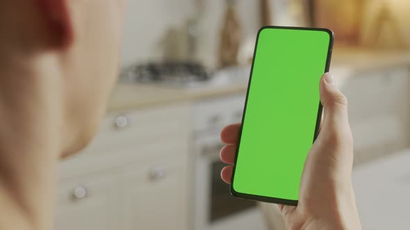 Back View of Man at Kitchen Room Sitting on a Chair Using Phone With Green Mockup Screen Chroma Key alt