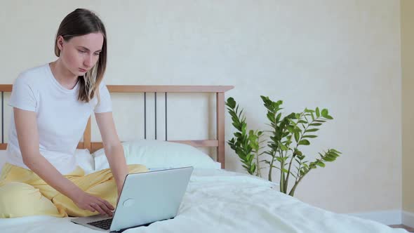 Woman Working on a Laptop Sitting on the Bed in the House. alt