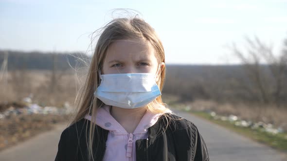 Portrait of Little Girl with Medical Face Mask Standing Outdoor. Sad Female Child Wearing Protective alt