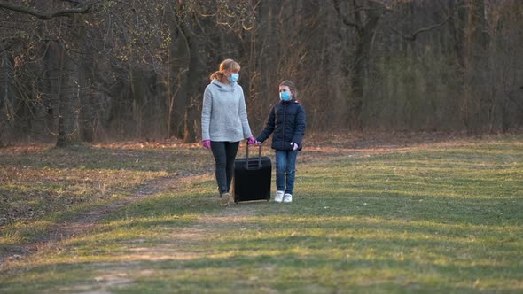 Mother and daughter walk through the spring Park in medical masks with a suitcase. 