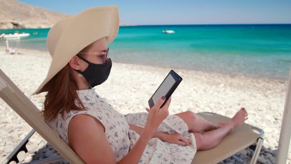 Woman in Face Mask and Hat Reads Ebook Relaxing on Sea Beach in Summer Day