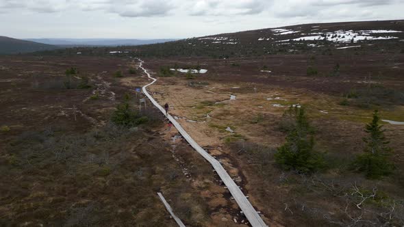 A woman and her dog walking on a mountain plank path with snow behind them. Fast aerial orbiting inf alt