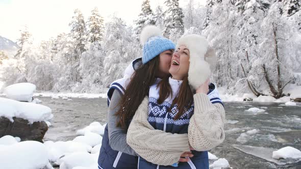 Two Happy Girls Laughing Hugging Each Other Near a River in a Snowy Forest