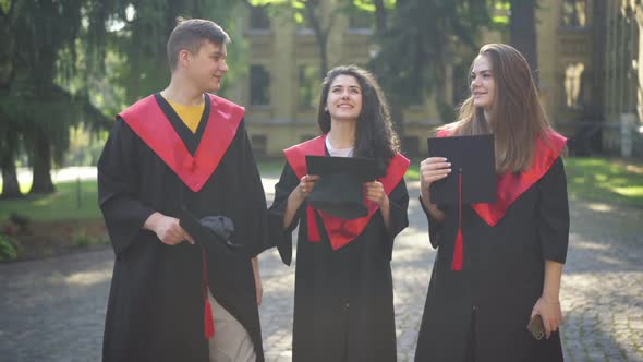 Front View Three Happy Students Throwing Mortarboard Cap on Sunny Morning Talking Smiling alt