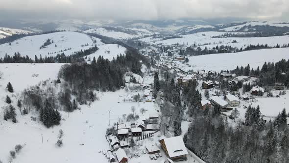 Aerial Winter Landscape with Small Rural Houses Between Snow Covered Forest in Cold Mountains