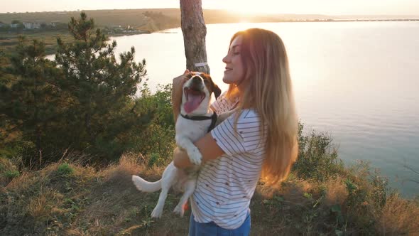Young Attractive Woman Playing with a Dog Jack Russell in the Meadow at Sunset with Sea Background alt