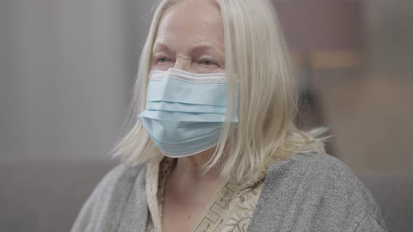 Closeup of Sad Lonely Old Woman in Covid19 Face Mask Indoors Turning to Camera alt