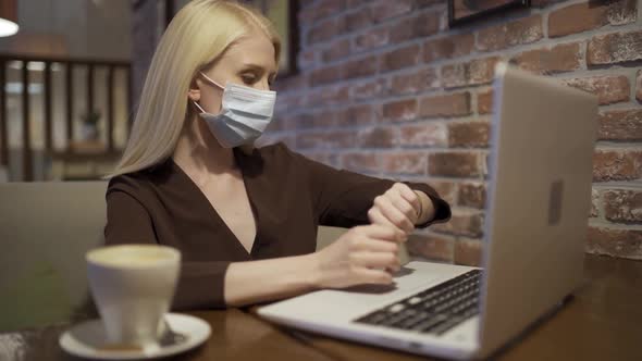 A Woman in a Medical Mask is Hastily Typing on a Laptop Keyboard and Looking at Her Watch in a Cafe