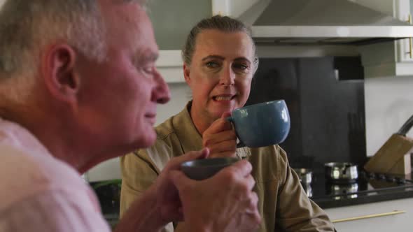Caucasian senior couple holding coffee cup talking to each other in the kitchen at home alt