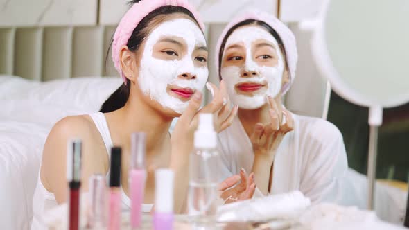 Beautiful young woman in white bathrobe applying a revitalizing  mask her friend's face. alt