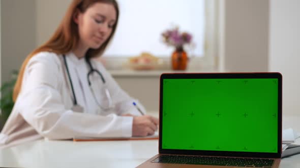 Laptop with Web Page Chromakey Mockup and Blurred Female Doctor Filling in Medical History at alt