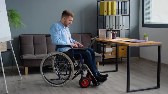 Disabled Person Freelancer is Working with Laptop Typing Sitting in Wheelchair at Office alt