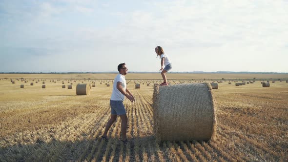 Little Daughter Jumping on Father's Arms From a Big Haycock in Field alt