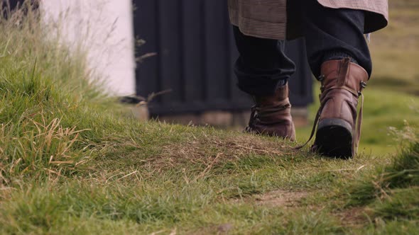 Mans Feet in Leather Boots Walks on Grass Towards Little Black House alt