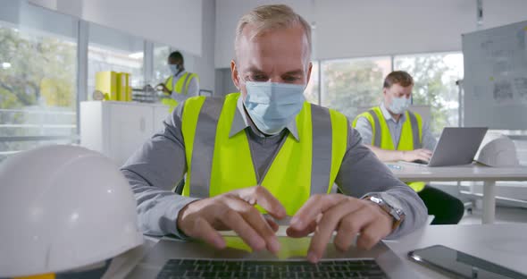Mature Architect in Face Mask Working on Laptop Sitting at Table in Office alt