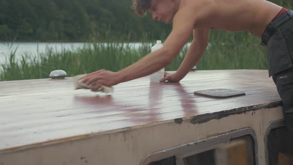 Young carpenter brushing down sanded roof planking of old wood boat using acetone on cloth in hot Su alt
