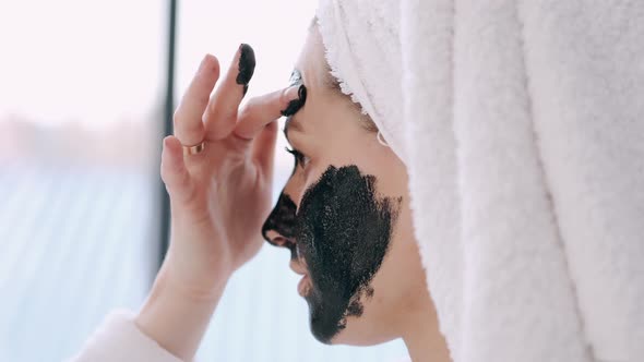 Pretty Woman with Wrapped Hair in a Towel Is Applying Black Mask on Face alt
